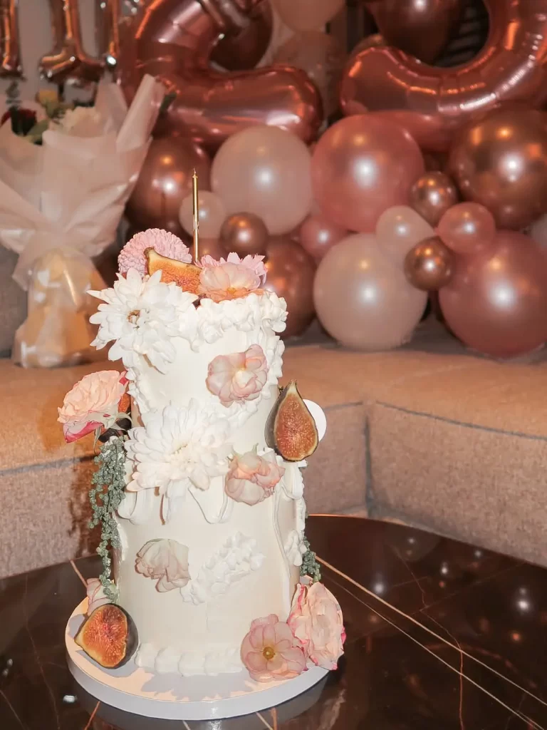 Tall white floral cake with pink blossoms and a single candle, against a backdrop of pink balloons.