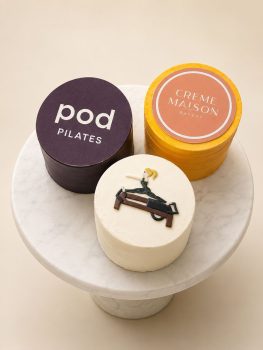 Three round bakery cakes on a marble stand: a dark purple cake labeled 'pod PILATES', a yellow cake labeled 'CREME MAISON BAKERY', and a white cake with a small sculpted figure sitting on a bench.