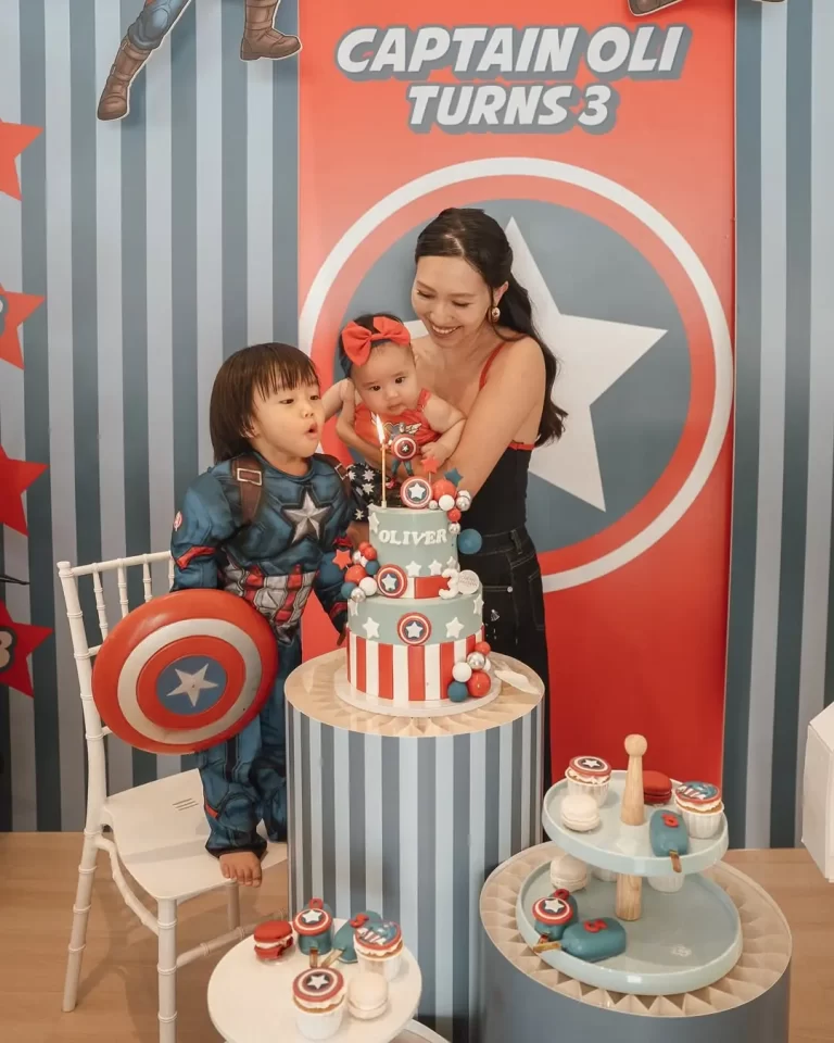 Mother holds a baby while a boy in a Captain America costume leans toward a tall patriotic birthday cake with candles and decorations behind them.