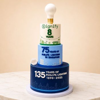 Four-tier cake commemorating Philips/Signify milestones: top white bulb, green-and-white eighth-year layer, blue '75 years' layer, and dark blue base with '135 years' message and skyline.