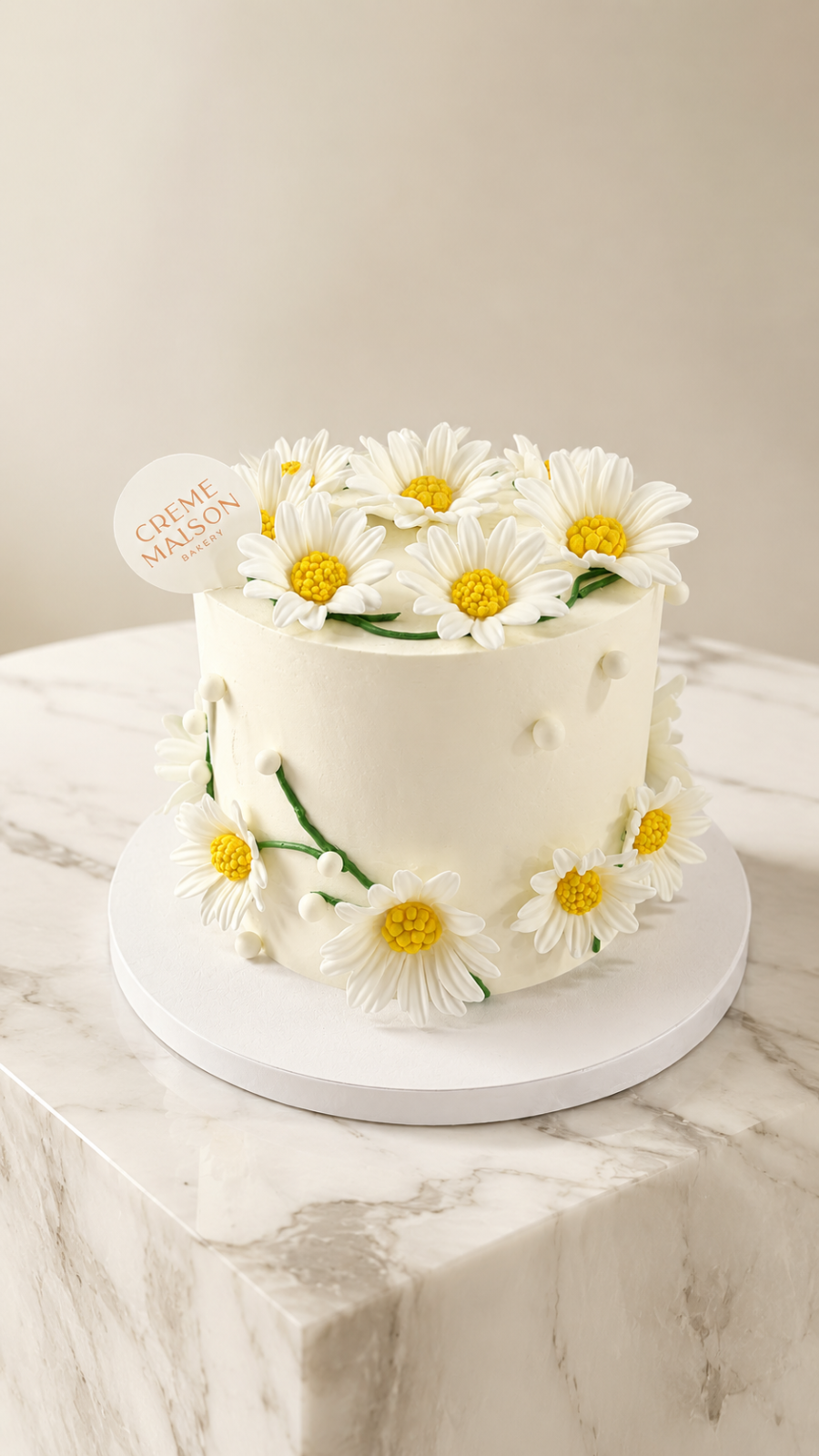White frosted cake topped and edged with white daisies and yellow centers, placed on a white cake board on a marble surface.