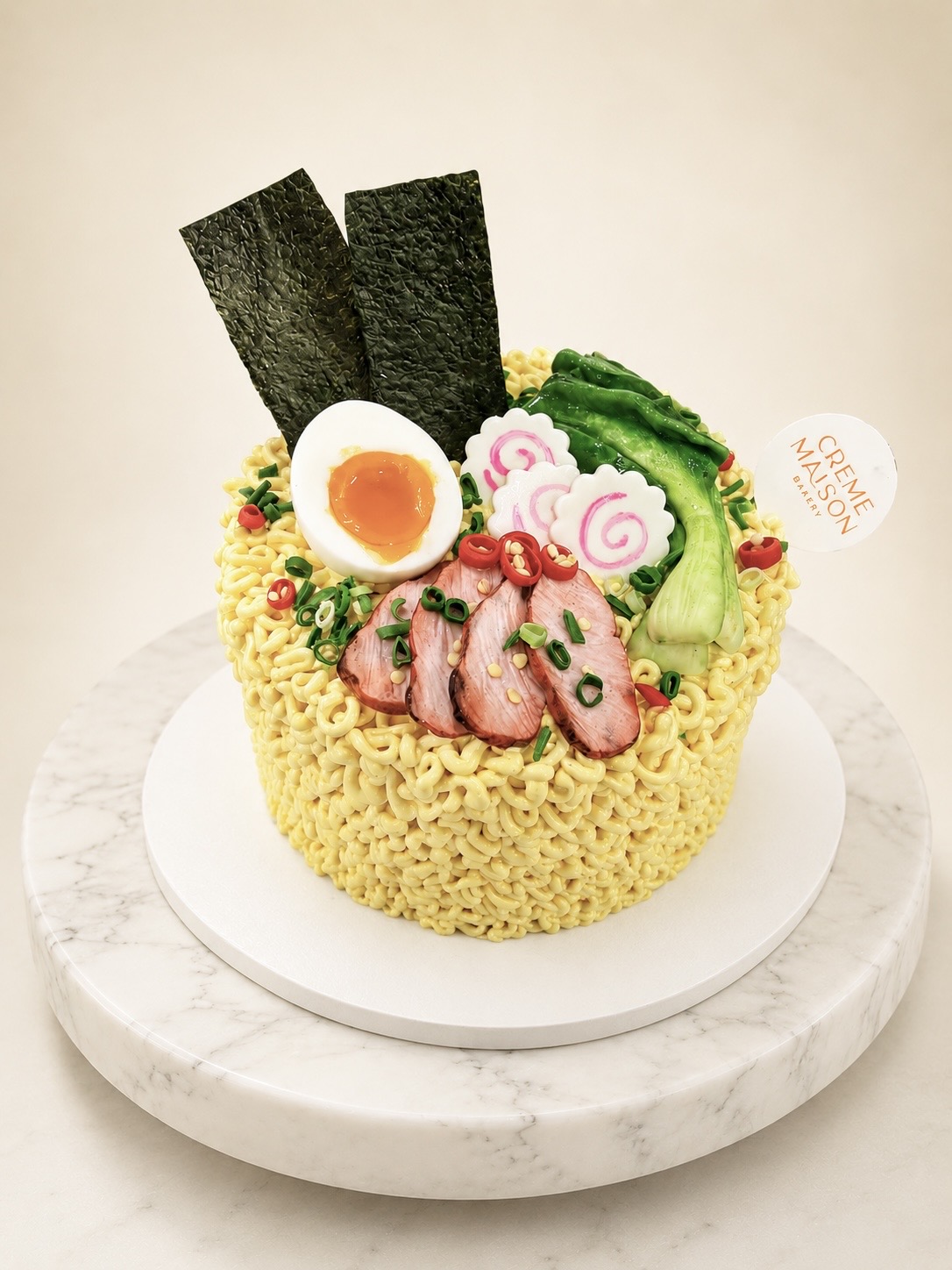 Ramen‑themed cake with yellow noodle frosting, pork slices, halved egg, nori sheets, narutomaki, and green onions on a marble board