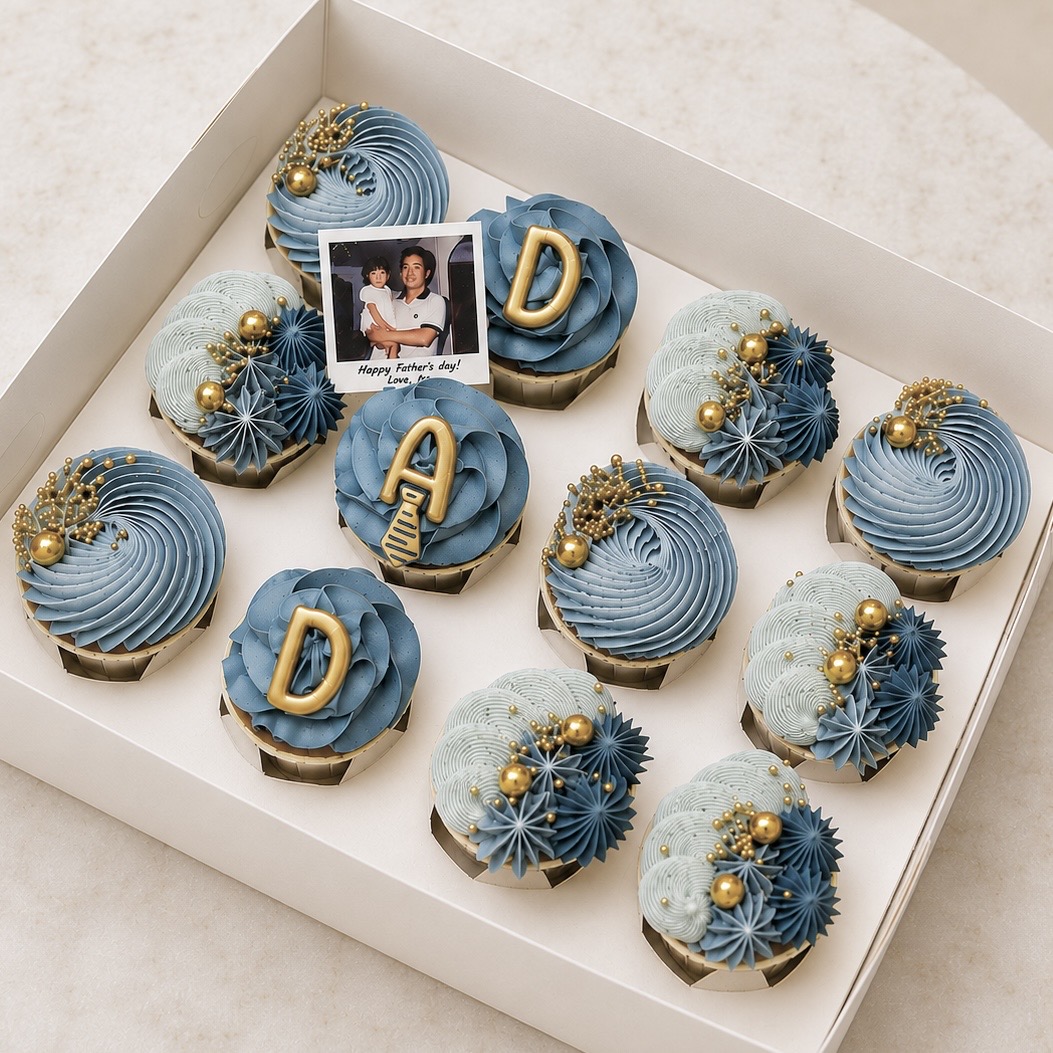 Box of blue-frosted cupcakes with gold bead decorations; some cupcakes have 'D' toppers and a small Father’s Day photo card in the box.