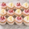 Cupcakes arranged in a grid on a marble surface, topped with white or pink swirls and strawberry halves with lemon zest optional.