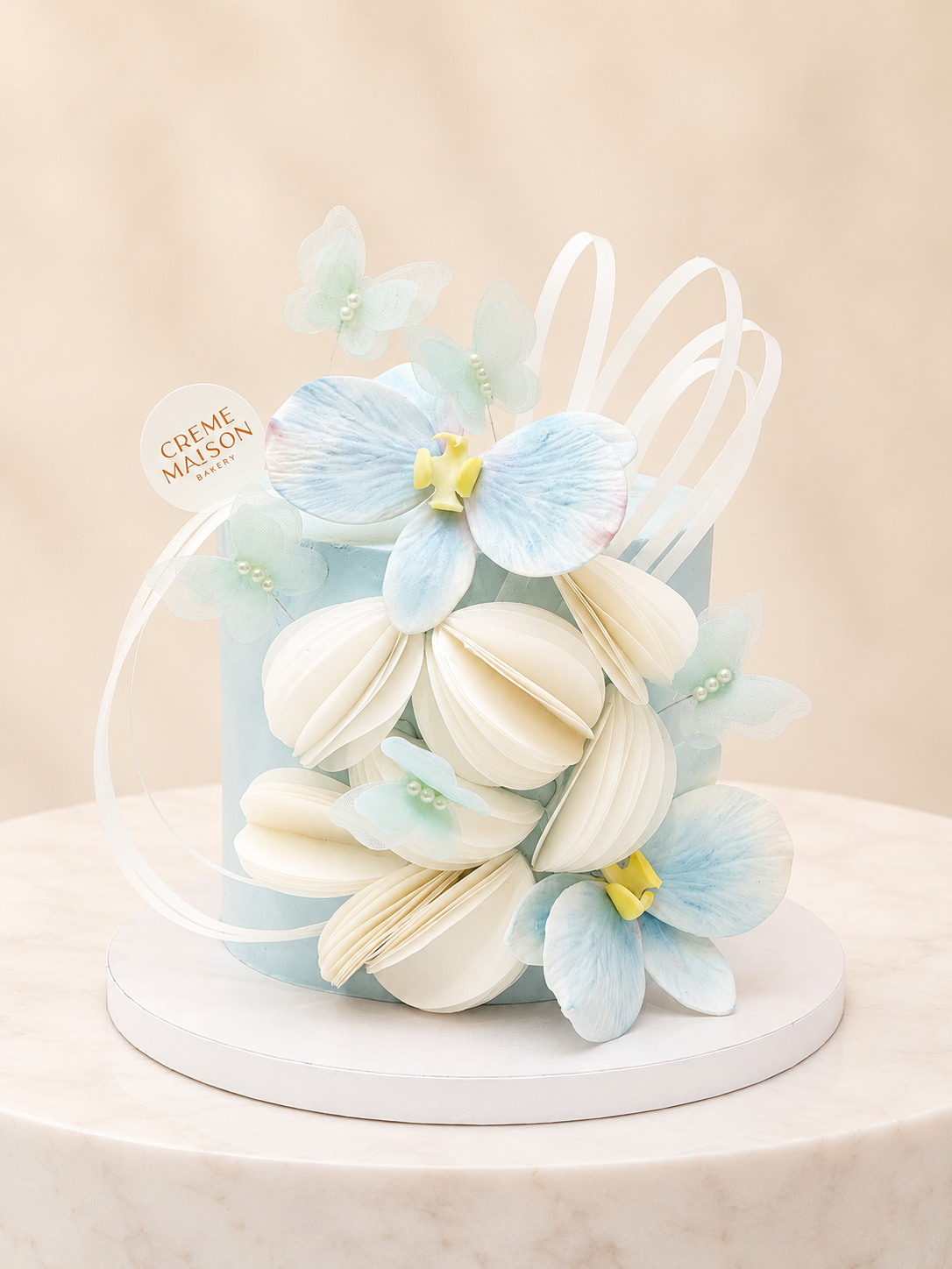 Pastel blue cake adorned with white sugar flowers, pale blue orchids, and curling white ribbons; Creme Maison Bakery tag visible.