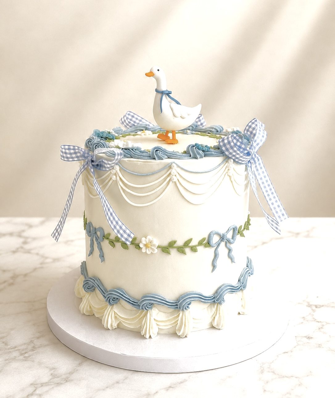 White fondant cake with blue swags, ribbon bows, and small white flowers topped by a white duck figurine wearing a blue scarf.
