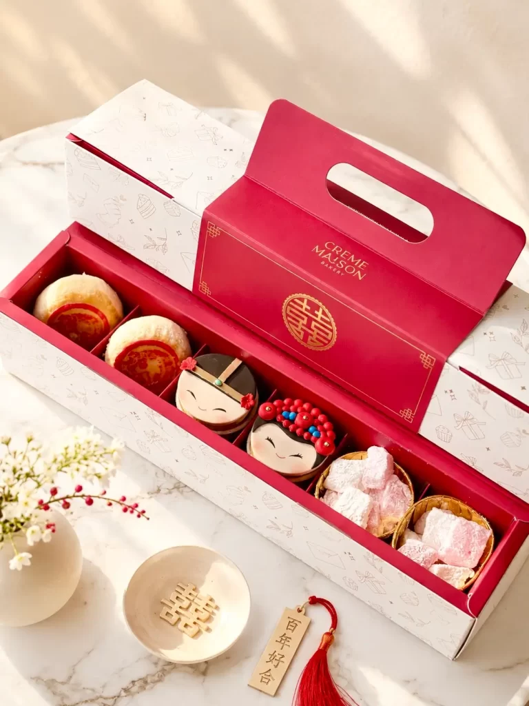 Open red gift box revealing six decorative pastries with cute face designs and pink/white sweets inside a white patterned outer box on a marble surface.