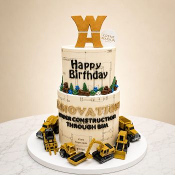Tiered birthday cake with a construction theme, featuring yellow toy excavators around the base and 'Happy Birthday' icing on the middle tier, topped with a gold logo topper and a small round 'Creme Maison Bakery' tag.