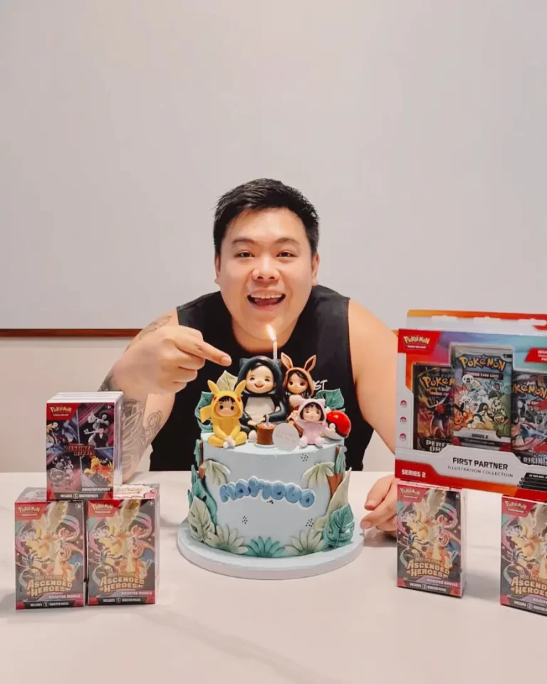 Person celebrating a birthday with a blue fondant cake topped with character figurines and a single lit candle, surrounded by Pokémon card boxes on a table.